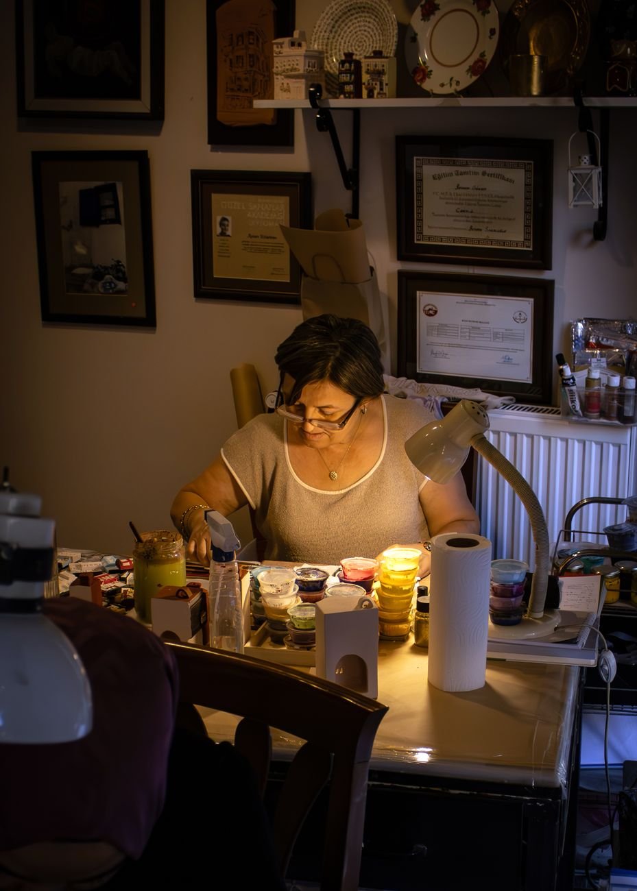 Woman artist paints under warm lamp light, surrounded by supplies in a cozy studio.