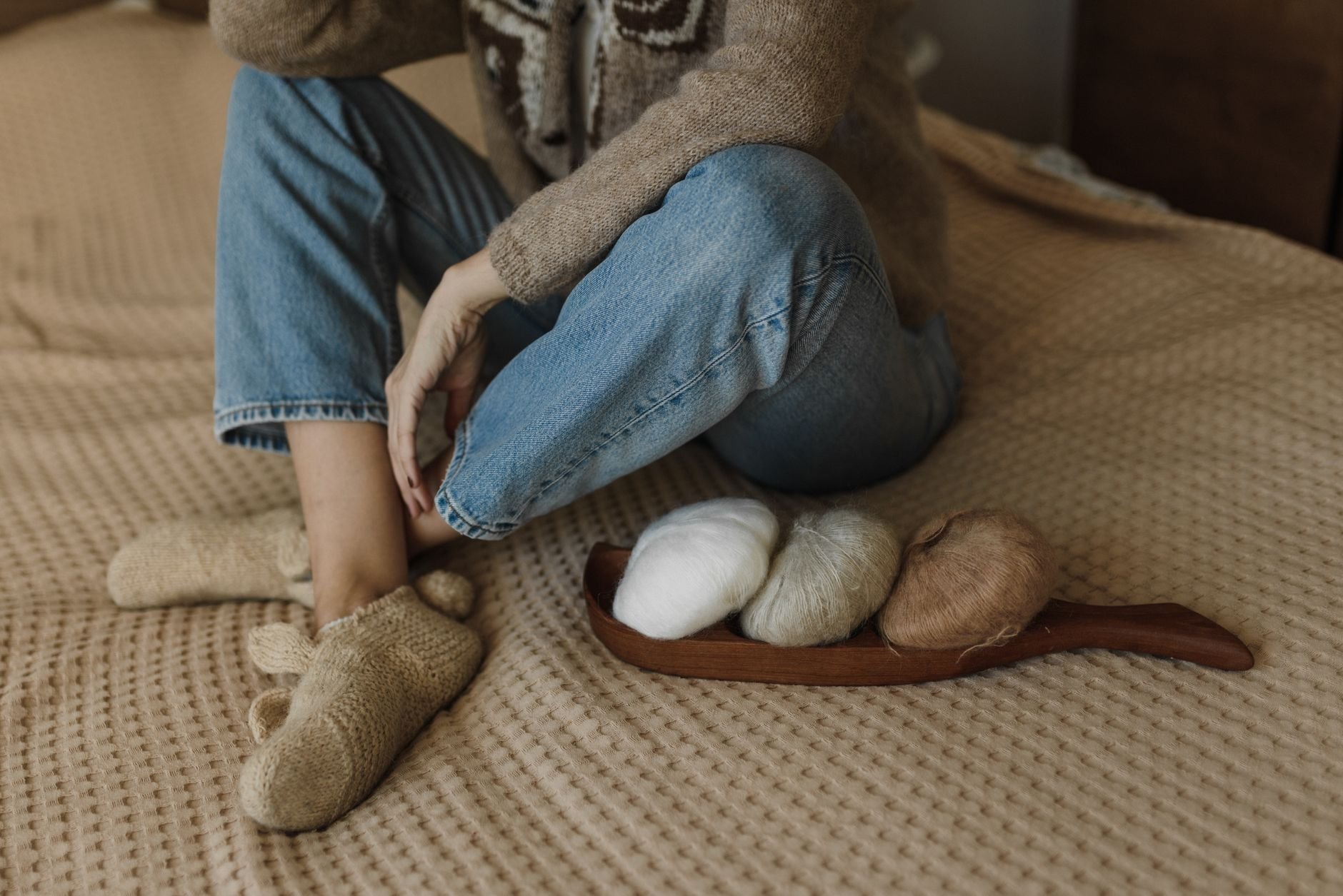 Woman sitting on a bed with yarn rolls, wearing cozy slippers, creating a warm crafting ambiance.